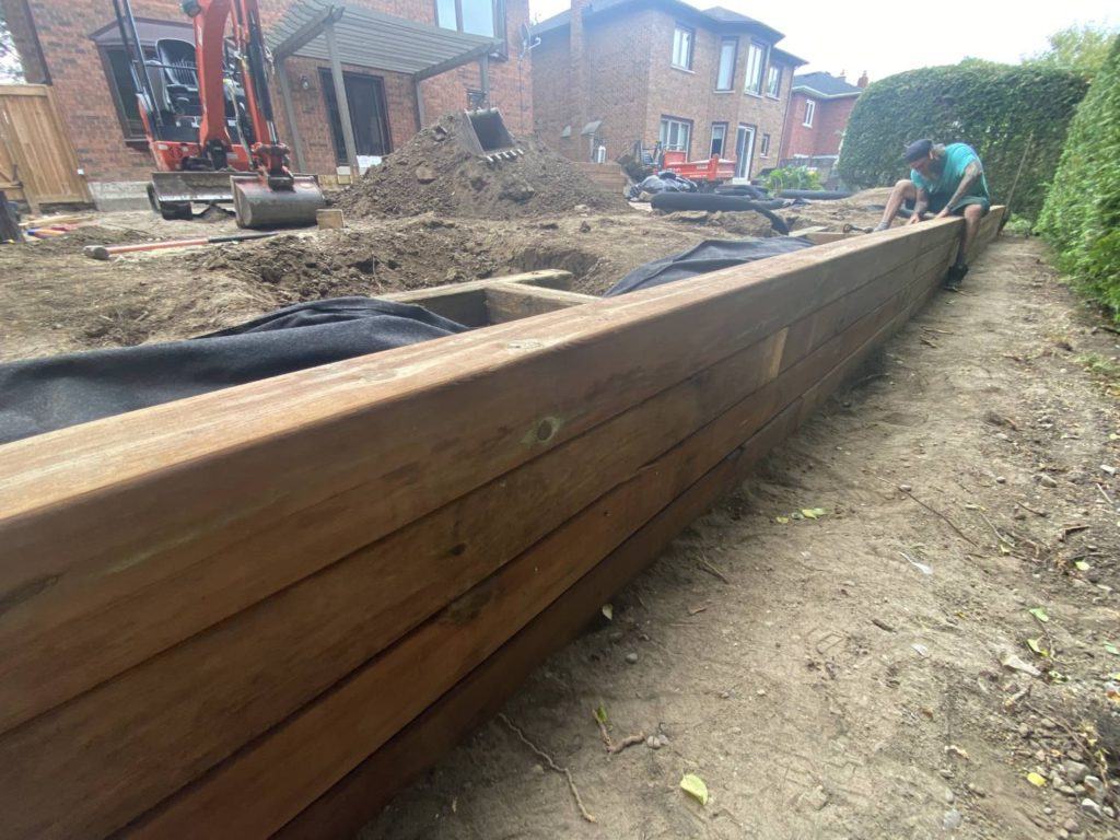 Timber retaining wall installation with excavator and backyard landscaping construction in progress