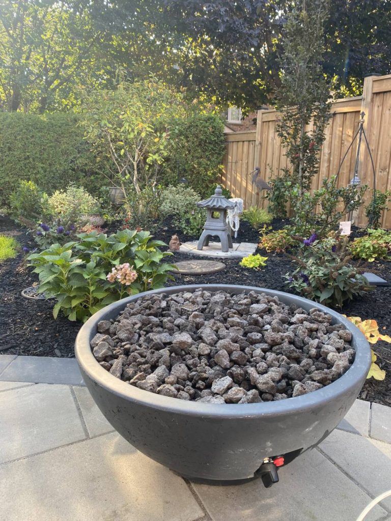 Outdoor fire bowl on stone patio beside landscaped backyard garden and seating area